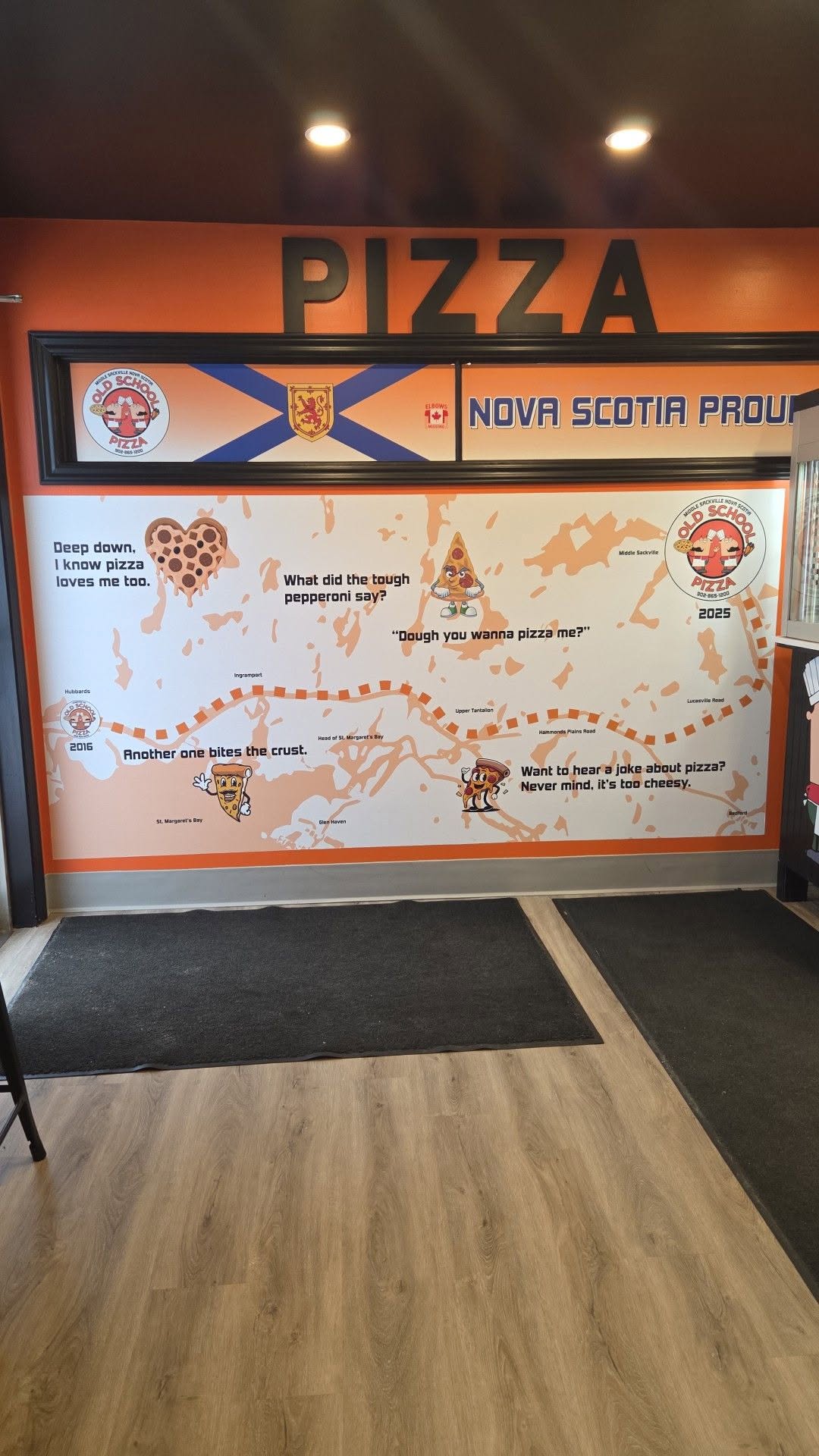 Inside Old School Pizza in Middle Sackville: an orange feature wall with the Nova Scotia flag, a hand-painted map of the South Shore and pizza jokes.